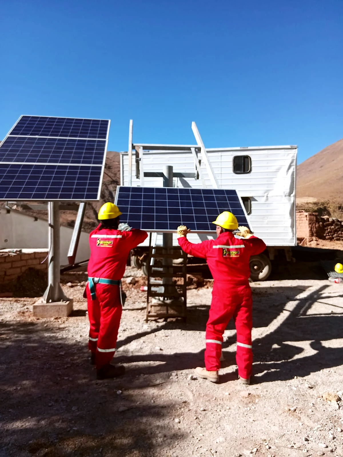 Dos operarios de Servicop con mameluco rojo y el logo en la espalda montan paneles solares en la montaña salteña